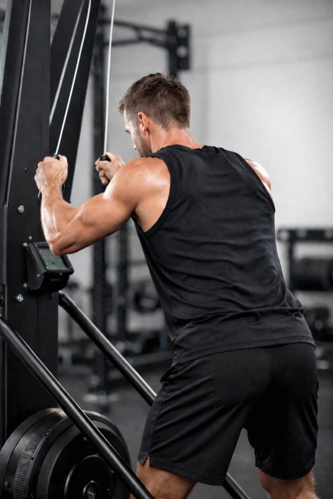 Back view of an athletic man using a SkiErg machine mid-pull, highlighting shoulder, arm, and core engagement in a modern gym.
