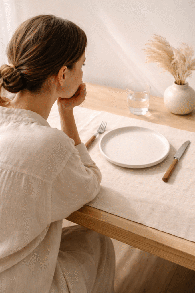 Person seated at a light wooden table, viewed from the side/back, gazing at an empty ceramic plate with fork and knife, soft natural light creating a calm, reflective minimalist dining atmosphere.