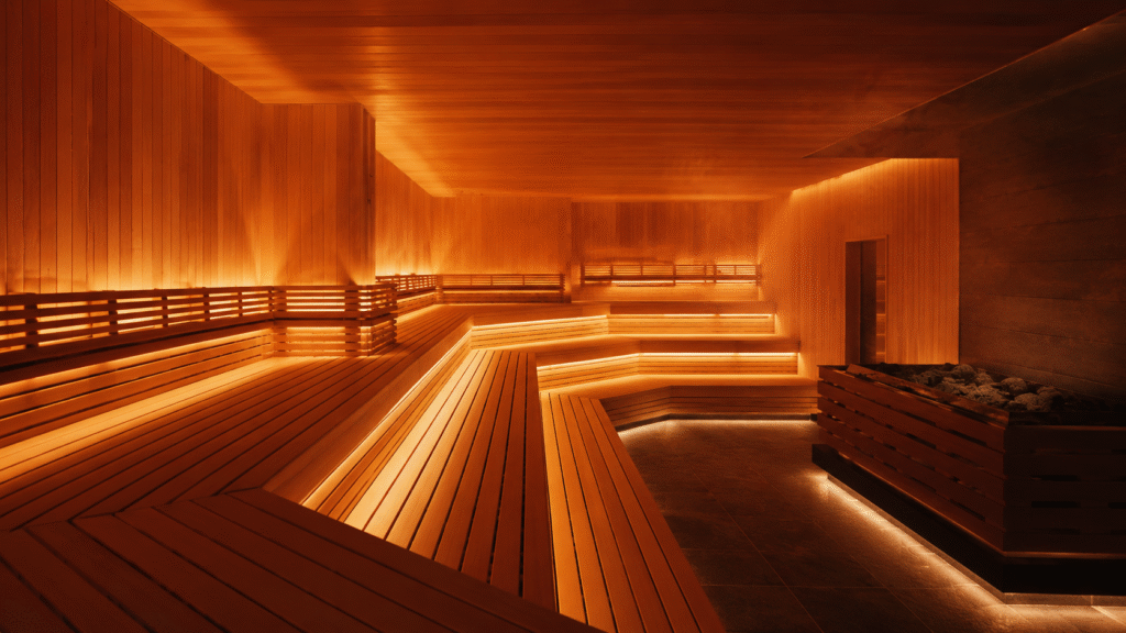 Spacious luxury sauna with tiered wooden benches, warm amber lighting, and a large stone heater.