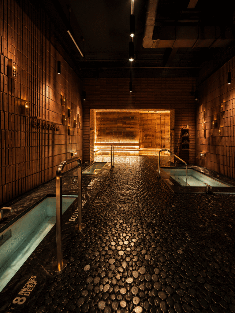 Cold plunge pools with metal handrails inside a dark, warmly lit wellness space with a pebble-stone floor.