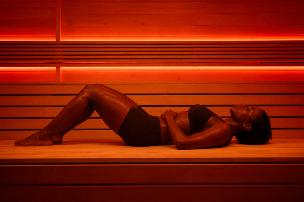 A woman lies on a wooden sauna bench with her eyes closed, relaxing beneath warm red-orange lighting.