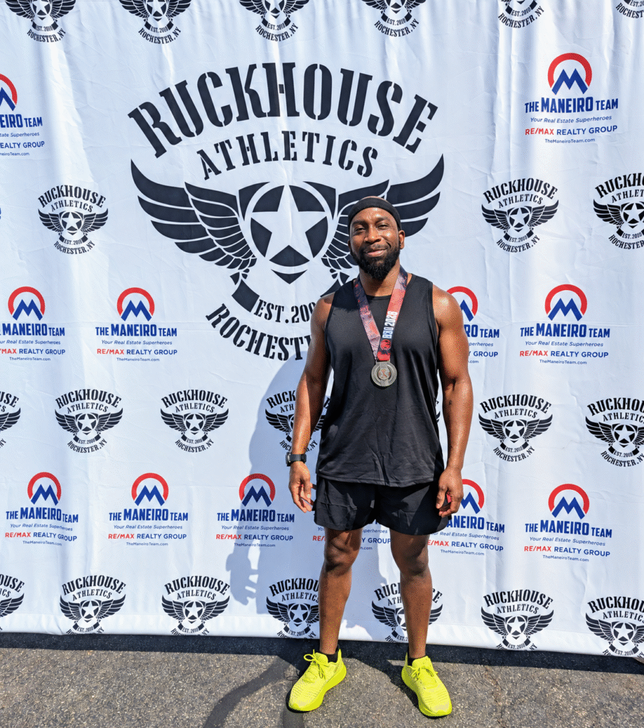 DEKA Mile finisher wearing a medal, black workout clothes, and neon yellow shoes in front of a Ruckhouse Athletics backdrop in Rochester, New York.