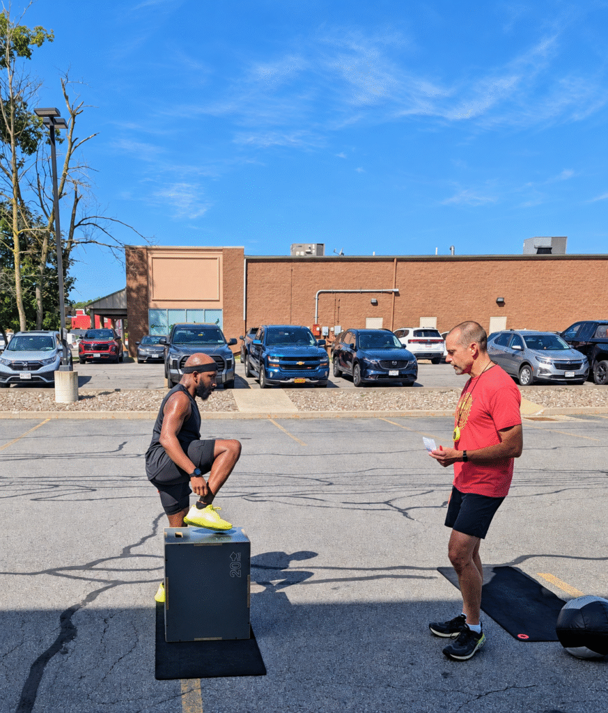 Athlete performing a box step-over during the DEKA Mile at Ruckhouse Athletics as a judge watches in an outdoor parking lot.