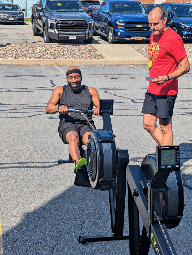 Athlete using a rowing machine outdoors during the DEKA Mile at Ruckhouse Athletics, with a judge standing beside him.