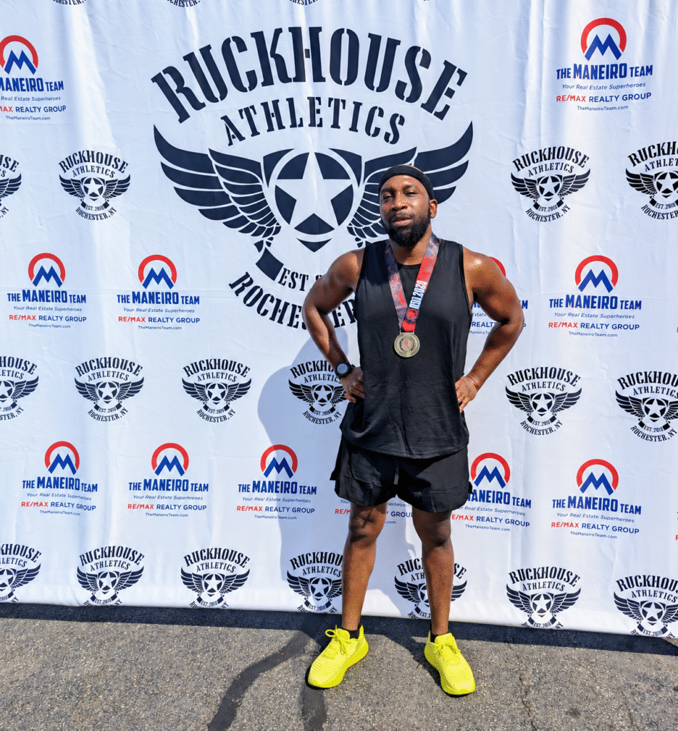 DEKA Mile finisher standing with hands on hips, wearing a medal in front of the Ruckhouse Athletics backdrop in Rochester, New York.